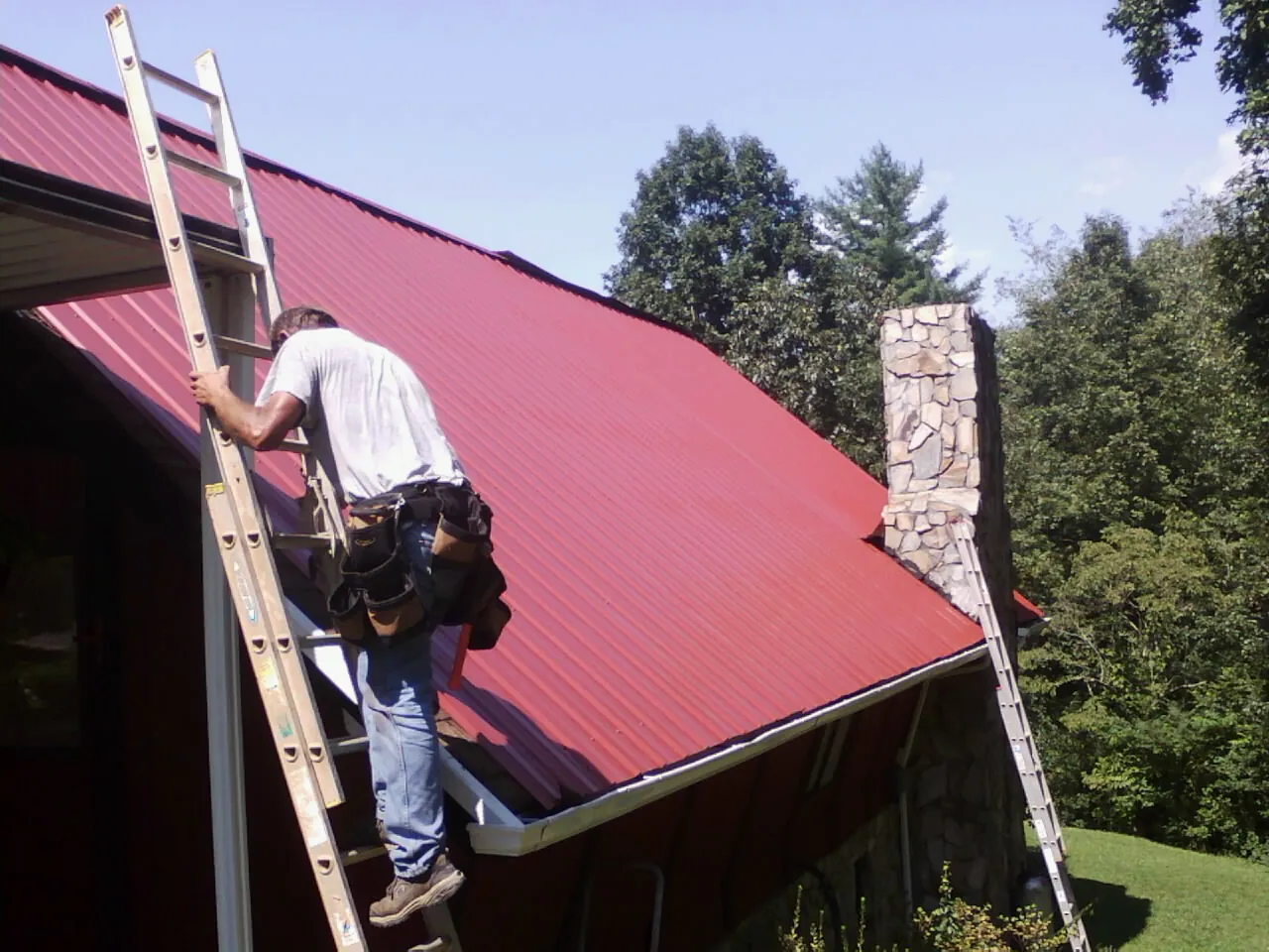 Roofer on a ladder beside a red metal roof and stone chimney — Soffit & Fascia Repair by Roofing Brothers in Saint Stephens Church